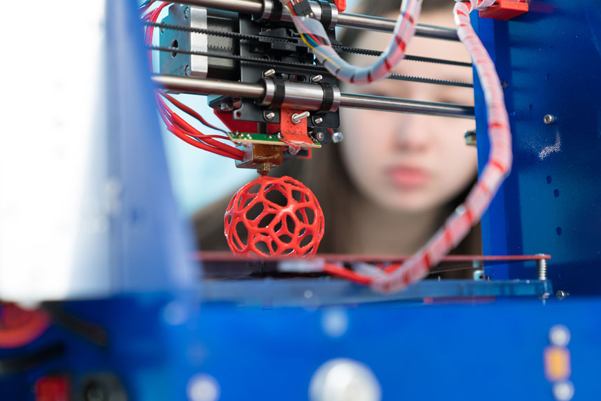 Một phụ nữ trẻ nhìn vào một máy in 3D tạo ra một nguyên mẫu hình quả bóng